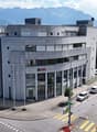 Large modern office building with concrete exterior, multiple floors, and parking spaces in front of the building. The building has the UBS logo displayed on the facade, indicating it is likely an office for the UBS financial services company. The surroun