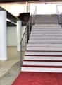The image shows a set of stairs with a red carpet leading up to the second floor. The stairs are made of granite or a similar stone material, with metal railings on both sides. The stairs are located in a large open space with white columns supporting the