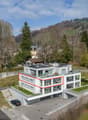 Modern white building, multiple floors, solar panels on the roof, balconies, parking area, surrounded by greenery