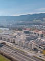 Aerial view of a city with a mix of residential and commercial buildings, highways, and parking lots. The city is surrounded by hills and mountains in the background, creating a scenic landscape.