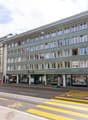 Five story building with green facade, multiple shops on the ground floor, tramway overhead.