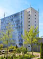 10 story white building, several windows, trees and bushes around, cars parked