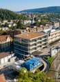 Aerial view of a cityscape with various buildings, trees, and a railway station in the foreground. The buildings are a mix of architectural styles, with some having red roofs. The trees are lush and green, and the railway station has several tracks and a