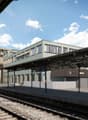 The image shows a modern train station with a covered platform and several buildings surrounding it. The station has a large, open design with glass walls and a metal roof structure. There are people waiting on the platform, and birds can be seen flying o