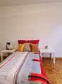 Spacious bedroom with hardwood flooring, white walls, a large bed with colorful bedding, side tables, a lamp, and a TV stand. The room also has a window, suggesting the presence of a balcony or terrace.