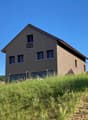 2-story wooden house with dark siding, large windows, and a balcony on the upper level. The house is situated on a grassy hill with trees in the background.