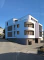 Modern white building, multiple balconies, large windows, parking area, mountains in the background