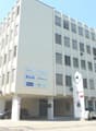 4-story office building, white exterior, many windows with shutters, ground floor with parking space, signboards, air conditioners