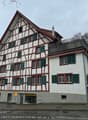 white and brown house, multiple windows, red and green shutters, brick chimney, balcony on top floor