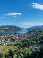 Scenic view of a lakeside town surrounded by mountains and lush greenery. The image shows a picturesque landscape with a large body of water, likely a lake, surrounded by hills and mountains. The town is situated along the shoreline, with colorful buildin