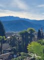 The image shows a scenic view of a mountainous landscape with a town nestled in the valley below. The foreground features lush greenery, including palm trees and other tropical vegetation, creating a picturesque and serene atmosphere. The background is do