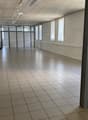 An empty, rectangular room with white walls, tiled flooring, and windows with blinds. The room is illuminated by white ceiling lights. It appears to be vacant with no furniture or decorations.