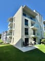 A three-story apartment building with modern design, balconies, and large windows, surrounded by a well-maintained lawn and lush greenery, creating a peaceful and picturesque environment.