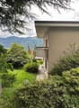 2-story house surrounded by lush greenery, including trees and shrubs. The house has a balcony or terrace overlooking the scenic mountain landscape in the background.
