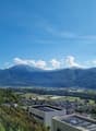 The image shows a panoramic view of a mountainous landscape with a town or village nestled in the valley below. The mountains in the background are tall and rugged, with a blue sky dotted with clouds overhead. The foreground includes some buildings or str