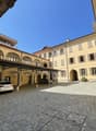 Cobblestone courtyard with cars parked under a covered area, yellow building with arched windows and doors, balconies, and a small plant in front of the entrance.