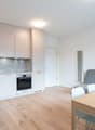 The image shows an open-plan kitchen and living room area. The kitchen features light-colored cabinets, a granite or stone backsplash, and a built-in oven and dishwasher. The living room area has a gray sofa and a wooden dining table with chairs. The floo