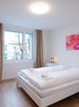 Spacious bedroom with white walls, large windows, hardwood flooring, a white bed with white bedding, two bedside tables with lamps, and a colorful abstract painting on the wall. The room also has a wooden ladder-like structure, possibly for storage or dec