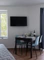 The image shows a studio apartment with a living/sleeping area, a dining area, and a balcony. The living area has a large window with a view of the outdoors, a gray quilted bedspread, and a mounted TV. The dining area has a dark wooden table with blue cha