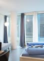 Spacious bedroom with large windows, gray sofa, laptop on a table, and a bed with blue bedding