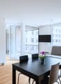 Spacious open-plan living area with large windows, hardwood floors, black dining table and chairs, gray sofa, built-in wardrobe, and access to a balcony or terrace with a view of the surrounding buildings.