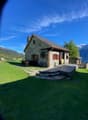 Single story stone house, red shutters, slate roof, porch, wooden wheel, located in a mountainous area with a view of the peaks