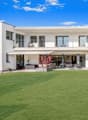 Two-story modern white house with large windows, balcony, and patio area overlooking a well-manicured lawn and garden. The house has a clean, minimalist design with a flat roof and appears to be situated in a scenic, mountainous area with a clear blue sky