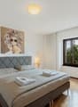Spacious bedroom with a large bed, gray upholstered headboard, artwork on the wall, and a window overlooking the outdoors.