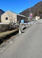 A single-story gray building with a slanted roof, surrounded by a stone wall and located in a mountainous area. There is a street with a speed limit sign visible in the foreground.