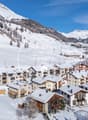 Snowy mountain landscape with a ski resort town nestled in the valley. The town features multi-story apartment buildings and chalets with balconies and parking spaces. The surrounding mountains are covered in snow, creating a picturesque winter scene.