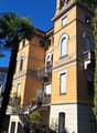 The building has multiple levels, each with balconies, a mix of beige and yellow wall colors, a tower-like section with distinct windows and arches, plants and trees around the area, and a shadow cast on the lower section.