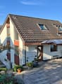 Detached house, brown roof, white walls, brown trim, two windows, two dormers, covered entrance, paved driveway, garden with plants and flowers
