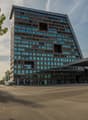 A large, modern office building with a glass and steel facade. The building has many floors and windows, and is surrounded by trees and a paved plaza area in front of the entrance.
