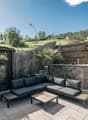 Outdoor patio with wooden deck, black patio furniture including a sectional sofa and coffee table, surrounded by a wooden fence and lush greenery. In the background, there is a castle tower visible on a hill.