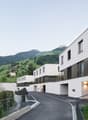 A modern, multi-story apartment building with balconies overlooking a scenic mountainous landscape. The building has a clean, minimalist design with large windows and a paved driveway leading to the entrance.