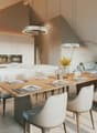 Dining room with a large wooden table, white chairs, a kitchen with modern appliances, and a living area with a couch and a window.