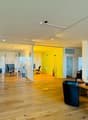 Empty office space with a glass wall, wooden floor, black office chairs, desk, brown wall, mounted picture frames, ceiling lamps.