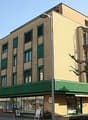 5 story building, brown walls, green roof, shops on ground floor, windows with shutters