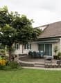 Two-story house with a tiled roof, surrounded by a well-maintained garden with a paved patio area and outdoor seating. The house has large windows and a covered balcony or terrace. There is also a parking area near the house.