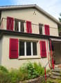 Two-story house with red shutters, beige walls, wooden terrace with a metal railing, awning over the entrance, and surrounded by greenery