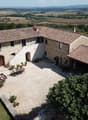 2-story stone farmhouse with red tile roof, courtyard with paved ground, outdoor seating area, surrounded by lush greenery and scenic countryside landscape