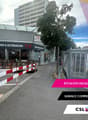 Facade of a commercial building with a sign reading 'ravaux intérieurs & extérieurs Tie', a closed shop with a canopy, a sidewalk, some barriers on the road, parked cars, trees and a building in the background.