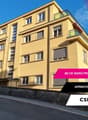 A multi-story yellow apartment building with balconies, located in Lausanne, featuring several windows, a driveway, and a bicycle parked in front.