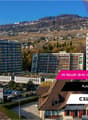 The image shows a multi-story building complex with several apartment buildings and a parking lot in the foreground. The buildings have a modern, residential design with balconies and large windows. The surrounding area appears to be a hilly, forested reg