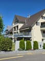 3-story residential building, beige exterior, brown roof, balconies on all floors, small garden, shrubs and trees in front
