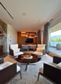 Modern living room with large windows, white chairs, black marble coffee table, and a TV on a wooden shelf.