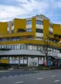 A yellow and white multi-story building with glass windows and balconies, located on a street with shops and trees.