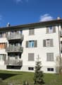 White apartment building with multiple balconies, brown roof, multiple windows, green grass in front