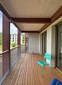 Wooden deck balcony with glass railing, overlooking a green, wooded area. The balcony has a turquoise chair and appears to be part of a larger residential building.