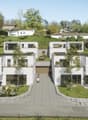 The image shows a modern residential complex with multiple two-story townhouse-style units. The buildings have a clean, minimalist design with large windows and balconies. The complex is surrounded by lush greenery, including trees and landscaped gardens.